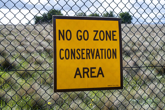 A bright yellow "No Go Zone Conservation Area" warning sign on a chain-link fence, protecting a native grassland habitat. Environmental preservation at&nbsp;Mt. Atkinson in Truganina, Australia.