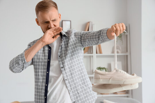 Foot odor and hygiene problem, man holding smelly shoes and covering nose with hand at home