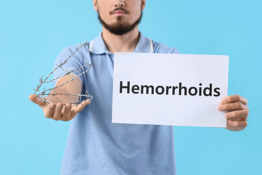 Young man holding paper with word HEMORRHOIDS and barbed wire on blue background, closeup