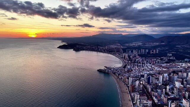 Aerial city bay under dramatic sunset clouds, glowing skyline and shimmering waterfront, moody orange light