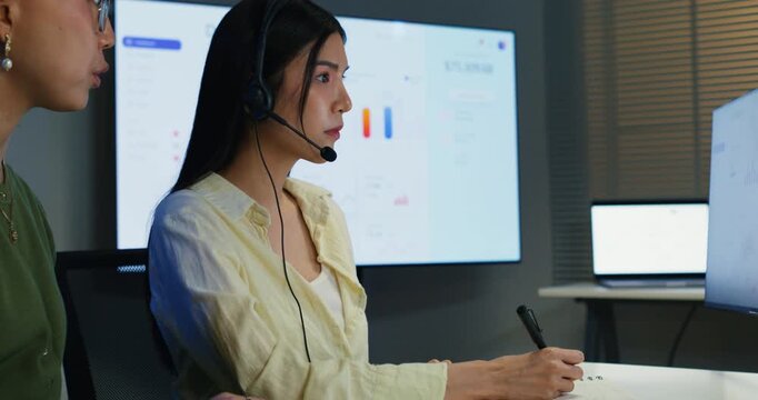 Asian call center team collaborating late at night, using headsets and computers to provide online customer service, demonstrating teamwork and dedication in a modern workplace.