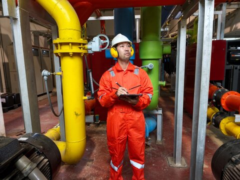An industrial engineer in an orange jumpsuit, safety helmet, and ear protection looking up while recording data on a digital tablet, surrounded by gas and cooling pipeline systems.