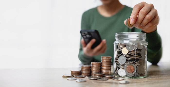 Woman putting coin into glass jar for saving money, with stacks of coins and smartphone on table. Financial planning and investment concept.