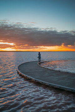 A silhouetted woman figure standing at the end of a sweeping curved jetty, looking across calm waters toward a vivid sunset, Lake Tyrrell, Victoria, Australia