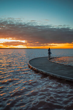 A silhouetted woman figure standing at the end of a sweeping curved jetty, looking across calm waters toward a vivid sunset, Lake Tyrrell, Victoria, Australia