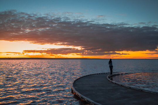 A silhouetted woman figure standing at the end of a sweeping curved jetty, looking across calm waters toward a vivid sunset, Lake Tyrrell, Victoria, Australia