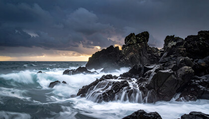 Fototapeta premium Dramatic seascape with dark, jagged rocks pounded by foamy waves under a stormy, cloudy sky. Sunlight subtly illuminates the horizon