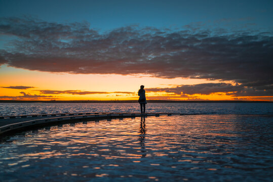 A silhouetted woman figure standing at the end of a sweeping curved jetty, looking across calm waters toward a vivid sunset, Lake Tyrrell, Victoria, Australia
