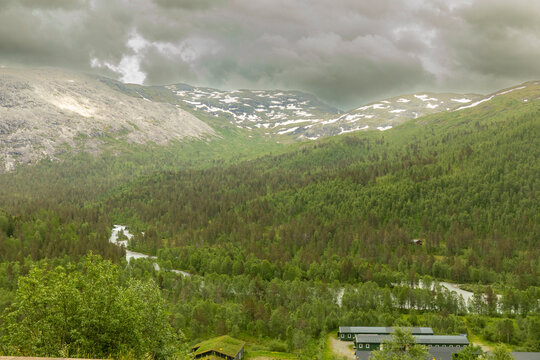 Part of the expansive Raundalen alpine valley in Norway, with a winding river cutting through dense pine forests beneath dramatic, cloud-covered mountain ridges and lingering snow patches.