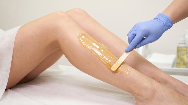 A professional beautician wearing blue rubber gloves applies warm liquid sugar paste or wax onto a woman's lower leg using a wooden spatula.
