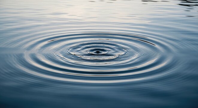 A single drop of water creates concentric ripples on a calm, dark blue body of water. The ripples spread outwards, distorting the surface of the water.