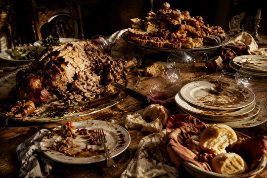Wide still-life of overloaded banquet table after long meal, torn roast, stacked plates, scattered bread, tipped wine glasses and pastries, gluttony mood