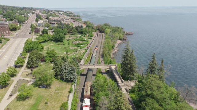 Aerial tracking shot follows train along Lake Superior shoreline past Leif Erikson Park Rose Garden bridge in Duluth Minnesota