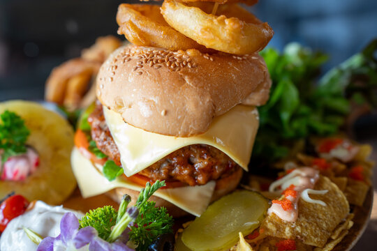 Delicious towering gourmet burger loaded with juicy meat, cheese, and stacked crispy onion rings on skewers, served with nachos and fresh salad in a restaurant with warm bokeh lights.
