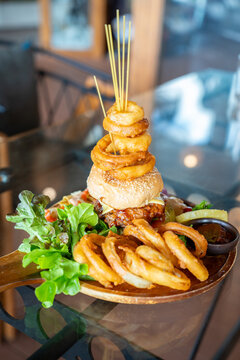 Delicious towering gourmet burger loaded with juicy meat, cheese, and stacked crispy onion rings on skewers, served with nachos and fresh salad in a restaurant with warm bokeh lights.