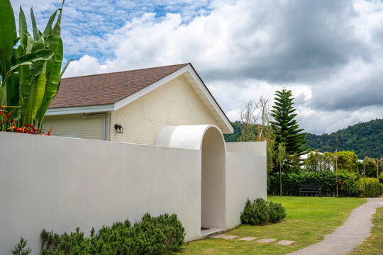 Modern white minimalist resort villa with an elegant archway entrance and solid wooden door, surrounded by tropical green plants and a white privacy wall under a bright blue cloudy sky.