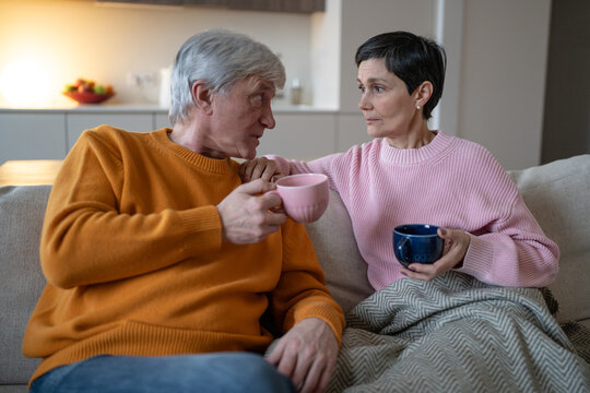Thoughtful wife listening to elderly husband while married couple relaxing together at home on sofa, senior man and woman talking and sharing thoughts, partners on couch in deep serious conversation.