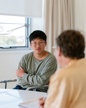 East Asian teenager sitting with a clinician in a dim room, featuring a guarded expression and glossy table for health services communication