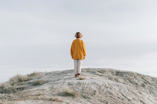 Child in a yellow sweater standing on a pale hill crest under a cloudy sky, featuring soft textures for solitude and wonder concepts