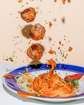 Meatballs falling toward a plate of spaghetti with glossy sauce and fresh herbs on a peach background, for food packaging and delivery advertising