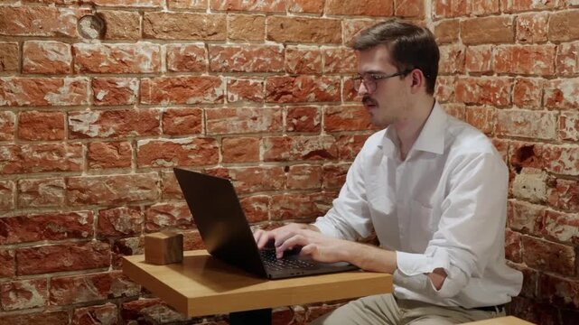 Young man in white shirt types on black laptop seated at wooden table against red brick wall. Consistent focus and posture across frames suggest steady work session in casual urban setting