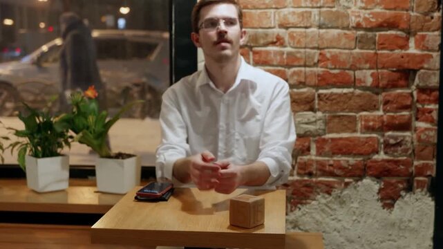 Video shows man entering frame, placing wooden cube and phone on table, then sitting down. Scene set in cafe with brick wall and window view. Man looks directly at camera