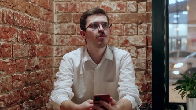 Man sits at wooden table against brick wall, starts thoughtful, then looks down and uses smartphone. Indoor scene with natural window light and green plant nearby