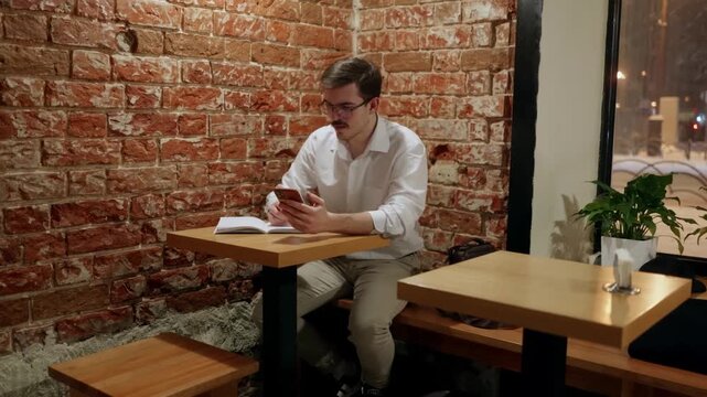 Young man in white shirt sits at wooden table against brick wall, focused on smartphone while notebook rests beside him