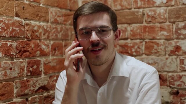 Young man in white shirt and glasses talks on phone against rustic brick wall. Expression shifts from focused to smiling. Indoor setting with warm lighting. Casual professional vibe