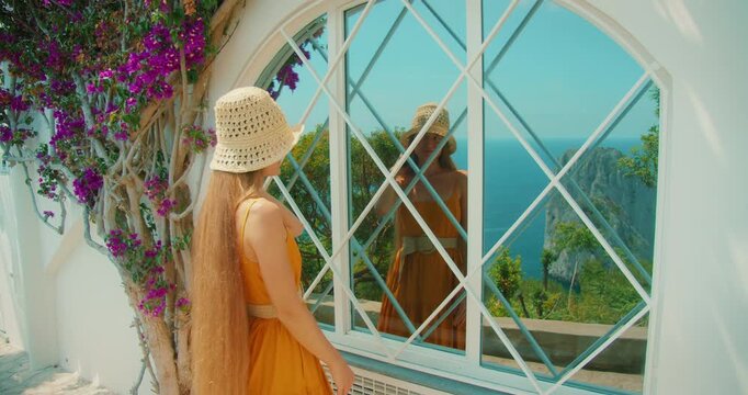 Woman visitor in summer dress admires her reflection by a window overlooking Capri cliffs on a bright summer day