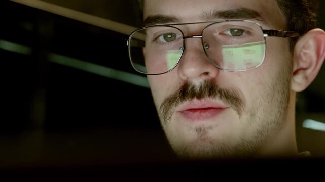 Close up of man wearing glasses with screen reflection visible in lenses. He appears concentrated, looking at computer monitor in dim environment