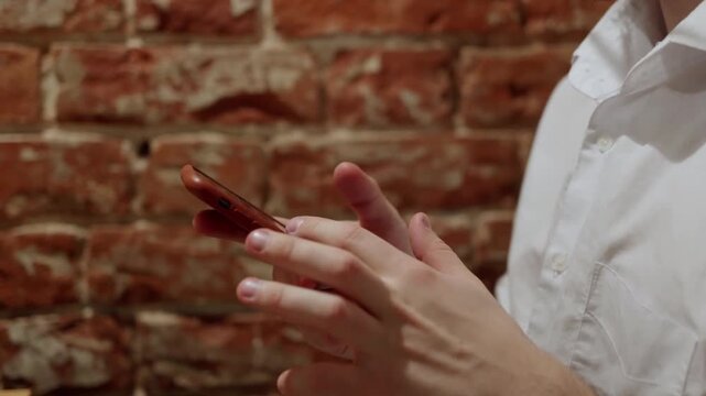 A man in white shirt interacts with red phone against rustic brick wall. Close up shows hands typing then looking up. Suggests focused communication or work in casual urban setting