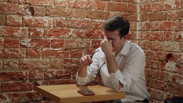 A man in white shirt sits at wooden table against brick wall. He rubs his eyes, adjusts his glasses, then looks up holding phone. Mood shifts from tired to alert
