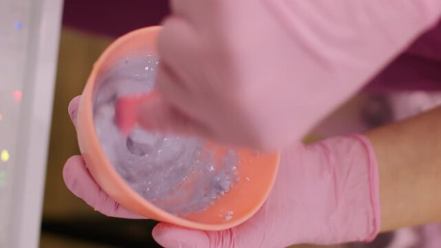 Vertical video. Mixing clay. Rejuvenation procedure. Unrecognizable female cosmetologist hands in pink protective latex gloves preparing mask product in orange bowl.