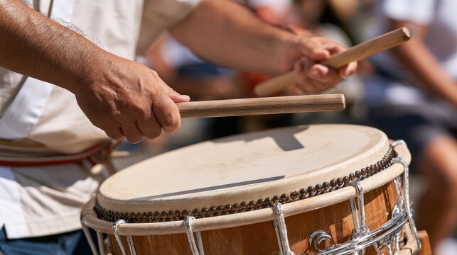 A close-up shot of a person's hands playing a drum with drumsticks. The drum is taut with a textured surface, and the hands are blurred with motion