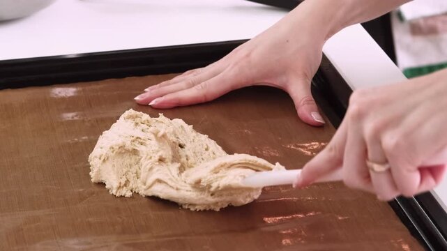 Preparing patisserie. Confectionery work. Female baker hand putting spreading dough with spatula on brown mat sheet on kitchen counter.
