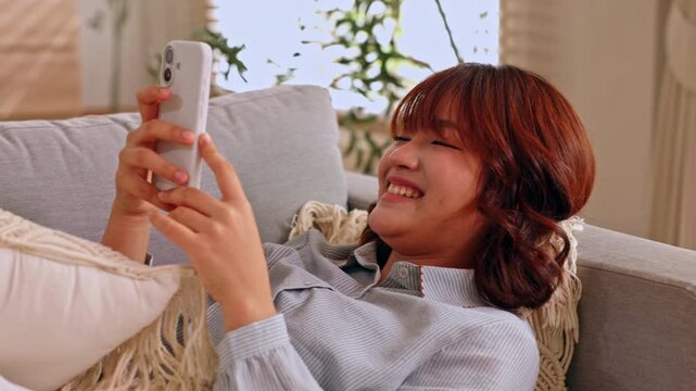 Young woman using smartphone on sofa with smile at home living room, red hair, casual relaxation, indoor texting, cozy couch, pillow, plant, soft daylight