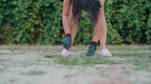 A nostalgic horizontal close-up shot of a gardener wearing protective gloves carefully pulling weeds from the paved walkway ground, filmed with a prime lens creating heavy background bokeh.