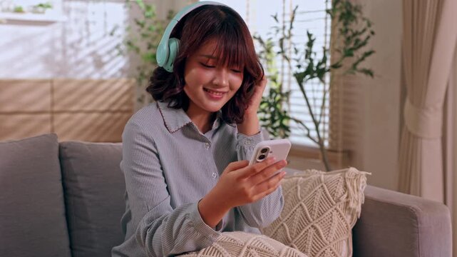 Young woman wearing headphone using smartphone on sofa in cozy living room, smiling while listening to music at home with casual relaxed lifestyle