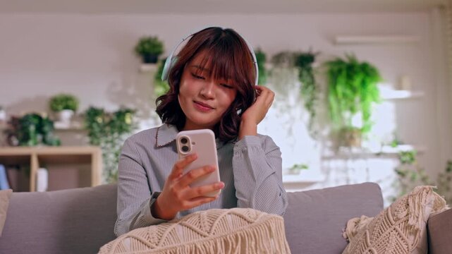 Young woman using phone on sofa in living room smile home plant message casual relaxed lifestyle listening to music cozy interior communication leisure theme