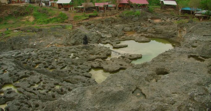 Tide pools formed in dark jagged volcanic rock formations near small local settlement at Broken Beach Nusa Penida Bali