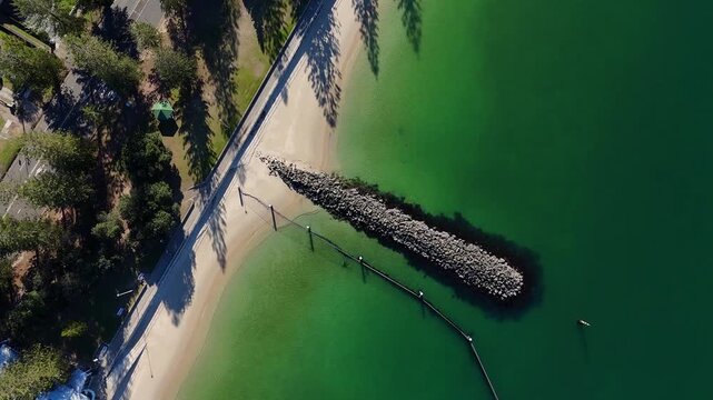Top down aerial of Ramsgate white sandy beach with tree shadows and rock groyne jutting rock pier breakwater and clear green water, drone ascend