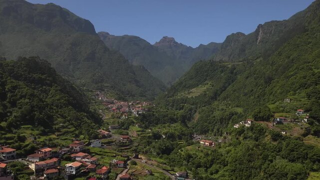 Aerial footage soars over Faja do Penedo, revealing red-roofed houses nestled in a lush, UNESCO-protected laurel forest valley beneath towering mountain peaks under clear blue skies.