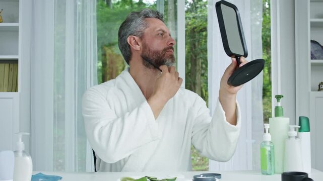 Man combing hair during morning routine at home. Bearded male grooming hair in front of mirror. Middle-aged man styling gray hair with comb. Male haircare and grooming concept.