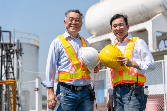 Smiling senior engineers holding hard hats at industrial plant, Mature factory managers in safety vests posing outdoors at facility, Professional industrial supervisors smiling at manufacturing site