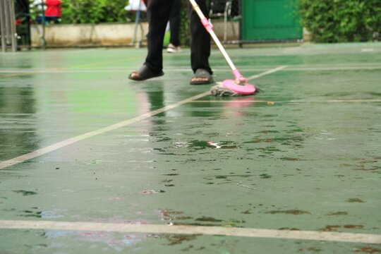 A maintenance worker mops a wet green sports court after rain, removing puddles and reflections for safety. The scene highlights routine cleaning, wet floor hazards, and outdoor facility upkeep.