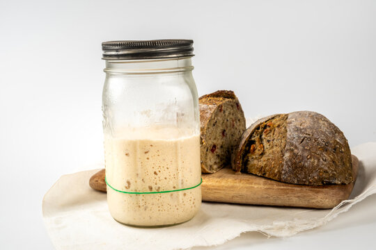 A glass jar filled with active starter in front of Crispy sourdough bread on a wooden board. Image on white background.