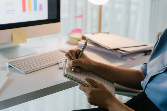 Person's hand writing in a spiral notebook, working at a computer desk with charts on screen, analyzing data