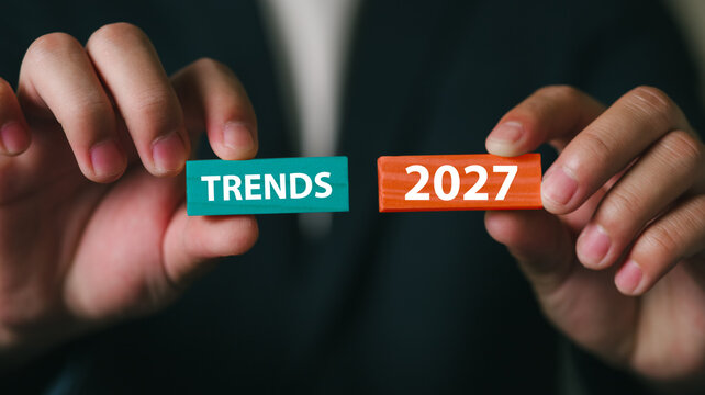 A professional in business attire holds blocks with the words Trends and 2027 in a modern office setting, symbolizing future planning, strategic thinking, and innovation.