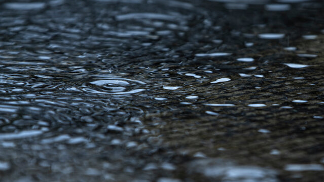 Close up of raindrops falling on water surface creating ripples during a storm.A moody and cinematic close-up shot capturing heavy raindrops falling onto a dark water surface